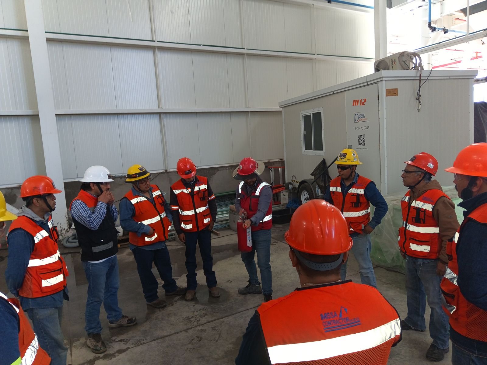 Cuadrilla con chalecos naranja y cascos reunida junto a una caseta de obra dentro de una nave industrial