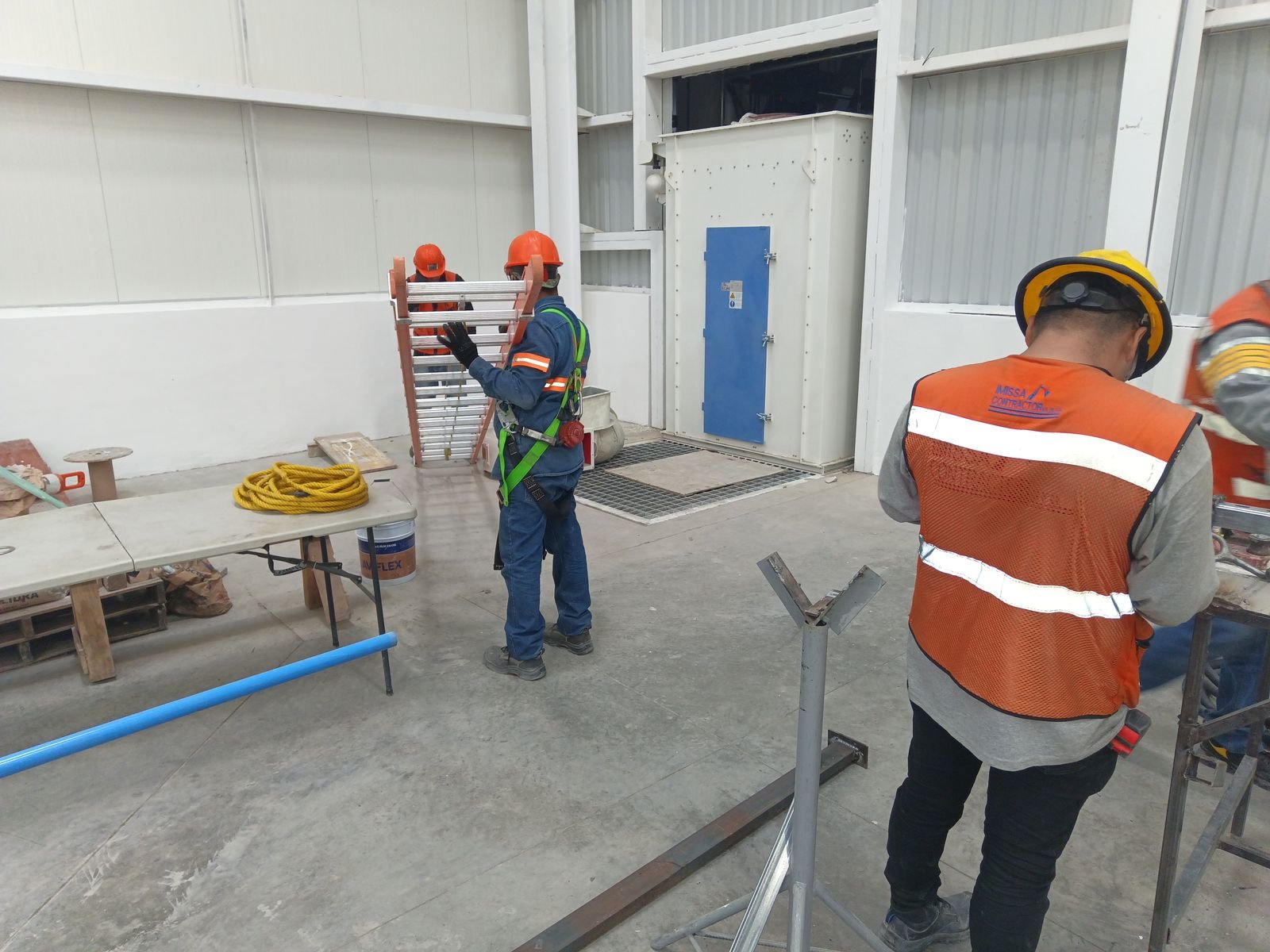 Trabajador con arnés de seguridad y compañero con chaleco naranja en un área de taller dentro de la nave
