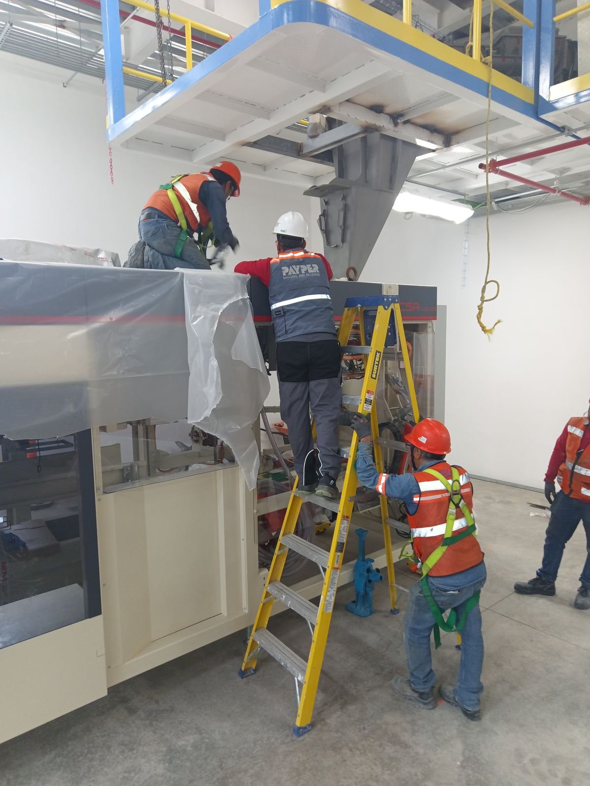 Técnicos con casco y arnés trabajando sobre escaleras en un equipo de empaque